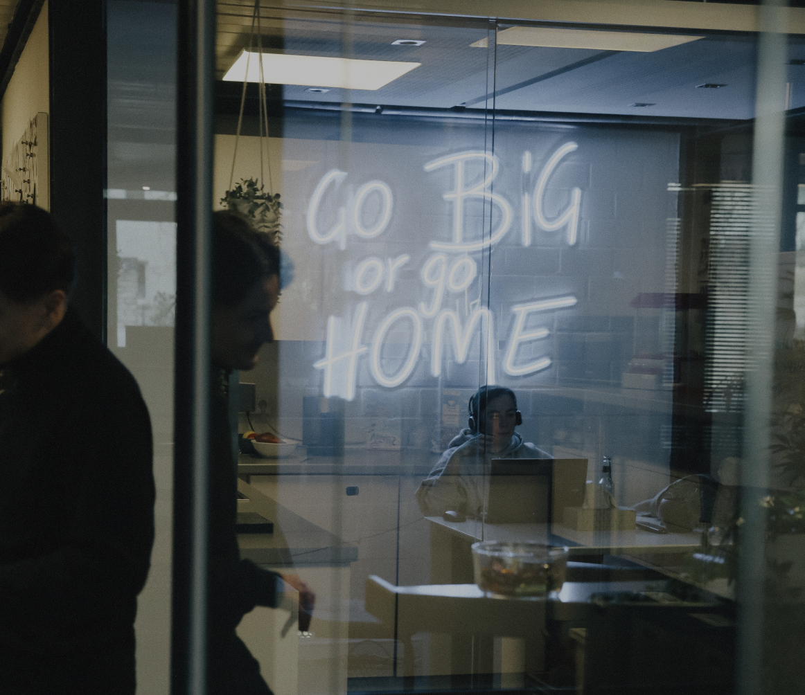Blick durch eine Glaswand in einen Besprechungsraum mit leuchtender Neon-Schrift „Go Big or Go Home“ und arbeitenden Personen