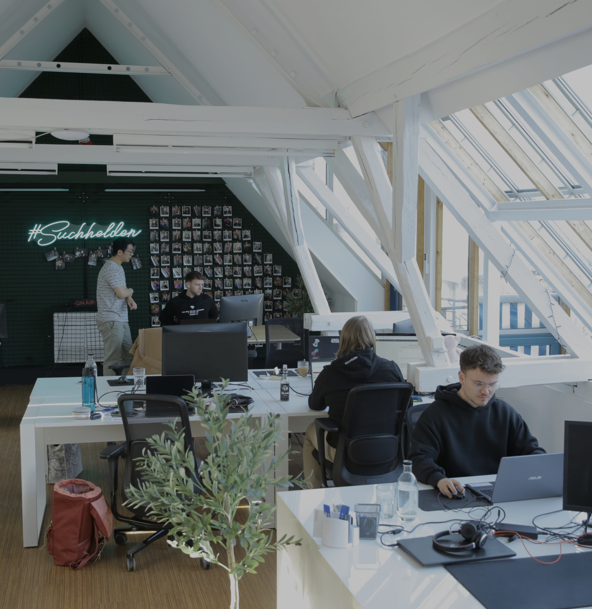 Mehrere Personen arbeiten an Schreibtischen in einem offenen Büro mit schrägen Dachfenstern, Pflanzen und einer beleuchteten „#Suchhelden“-Wand im Hintergrund