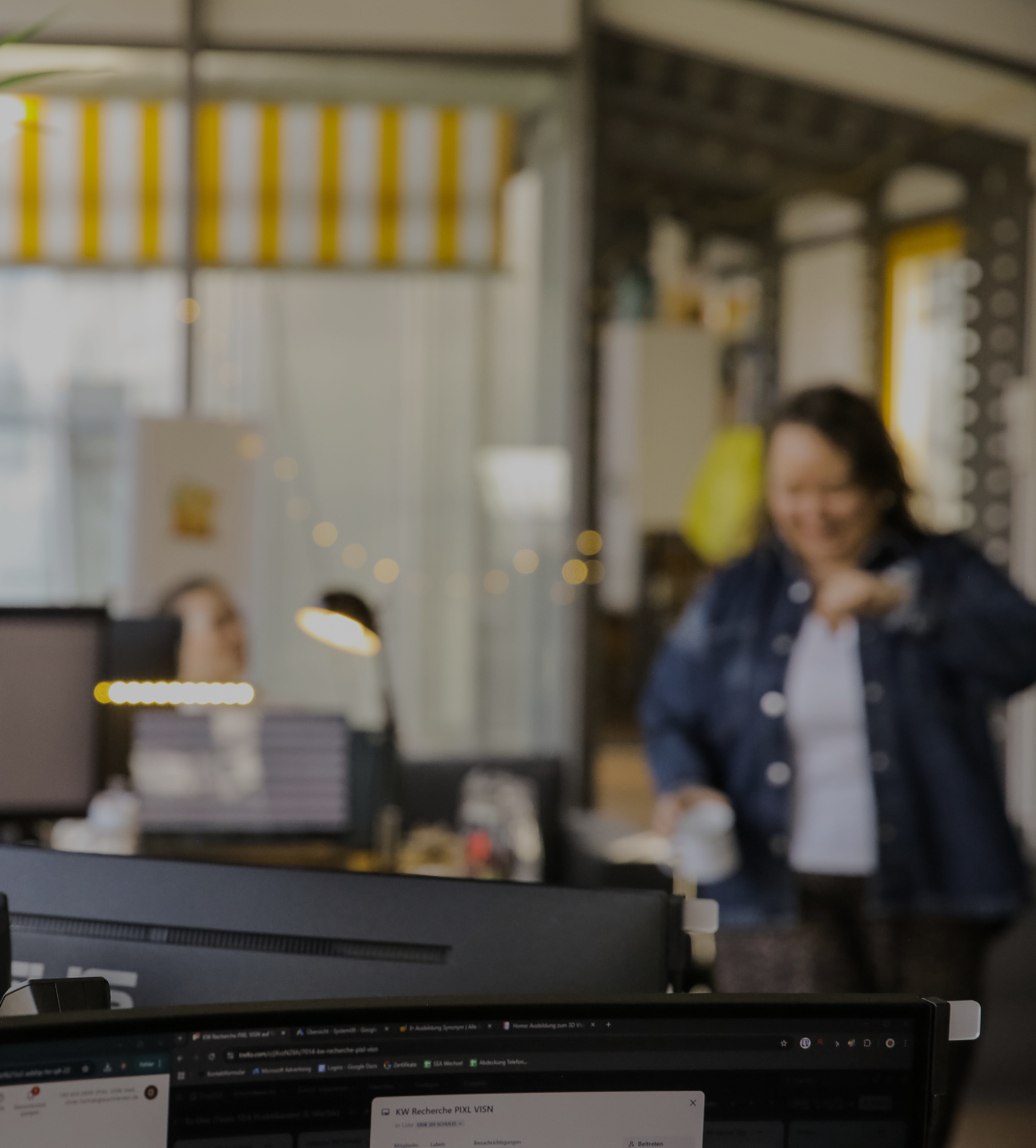 Unscharfe Büroszene mit Monitor im Vordergrund und einer Person, die im Hintergrund mit einer Tasse durch das Büro geht