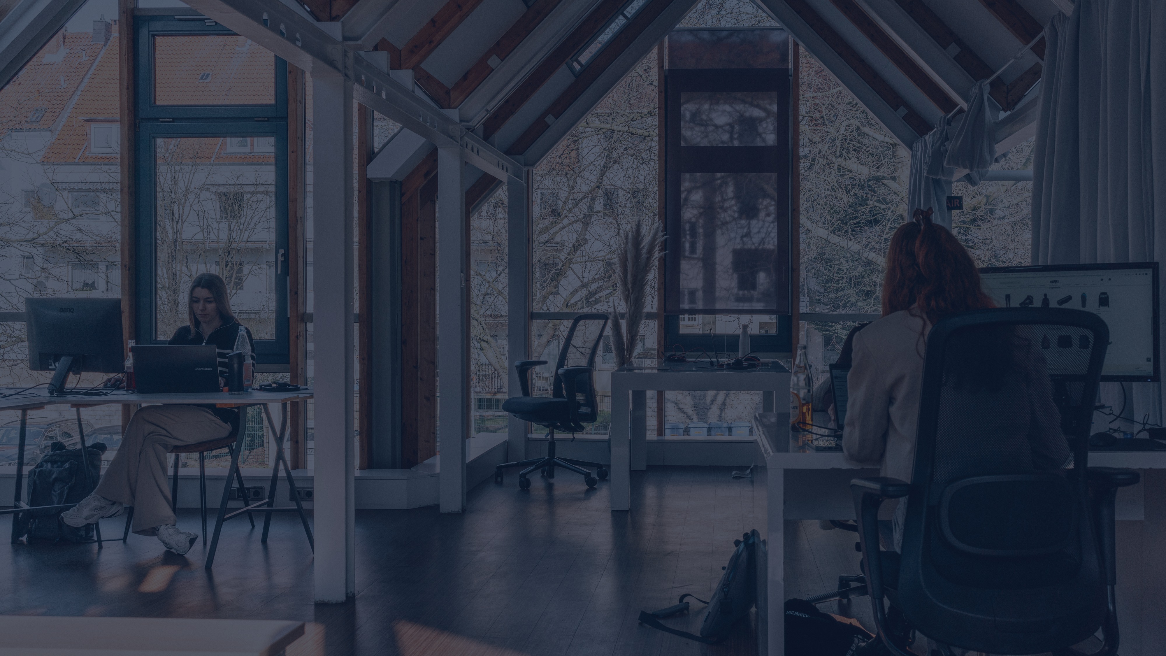 Zwei Personen arbeiten an separaten Schreibtischen in einem hellen Büro mit großen Fenstern, sichtbaren Holzbalken und Blick ins Grüne