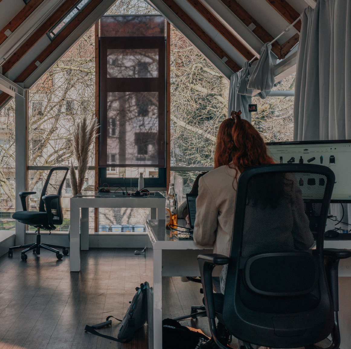 Zwei Personen arbeiten an separaten Schreibtischen in einem hellen Büro mit großen Fenstern, sichtbaren Holzbalken und Blick ins Grüne
