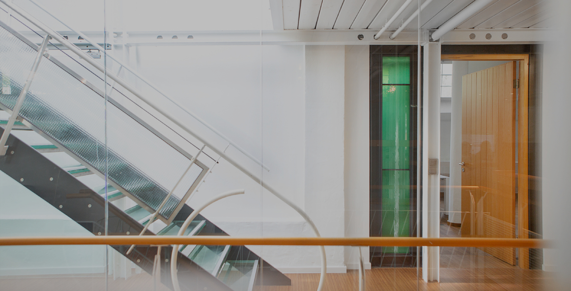 Heller Büroflur mit moderner Glas- und Metalltreppe, weißen Wänden und Holzboden; rechts ein offener Raum mit Holztür und grünem Glaselement, klare, minimalistische Architektur