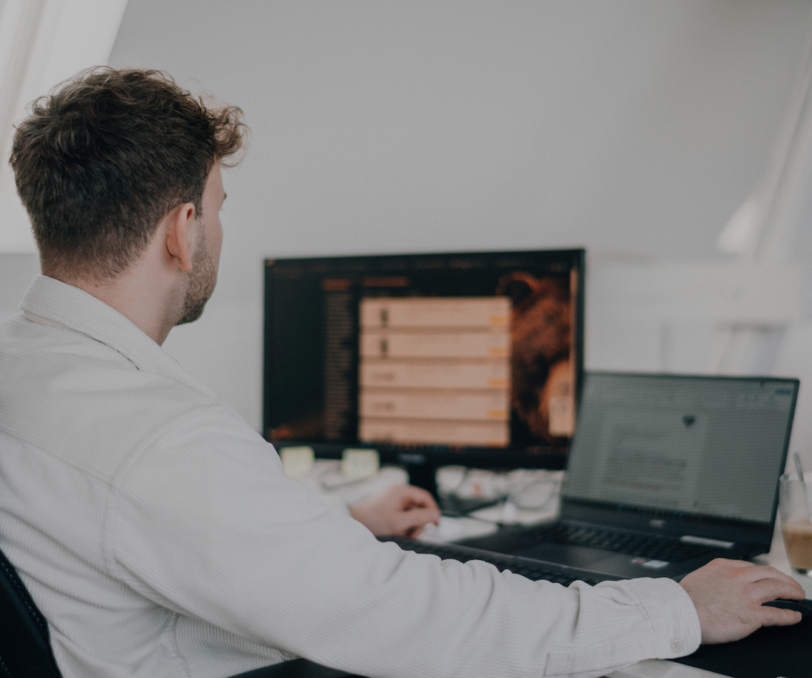 Person sitzt an einem Schreibtisch und arbeitet an einem Computer mit großem Monitor in einem hellen Büro, von hinten bzw. seitlich fotografiert