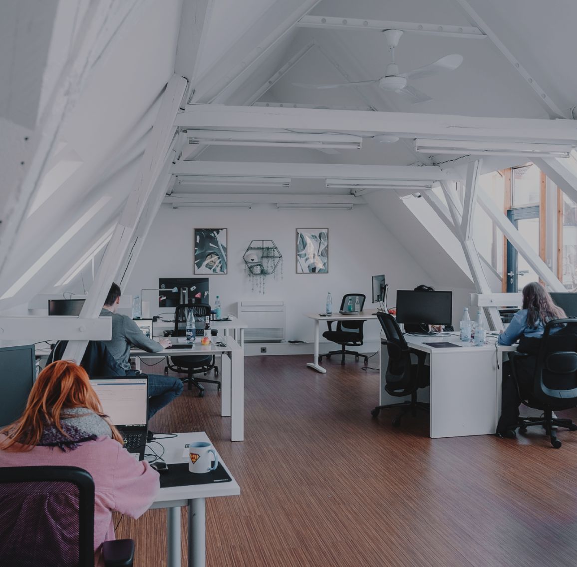 Großzügiges, helles Büro mit schrägen Dachbalken, mehreren Arbeitsplätzen und Personen, die an Schreibtischen arbeiten