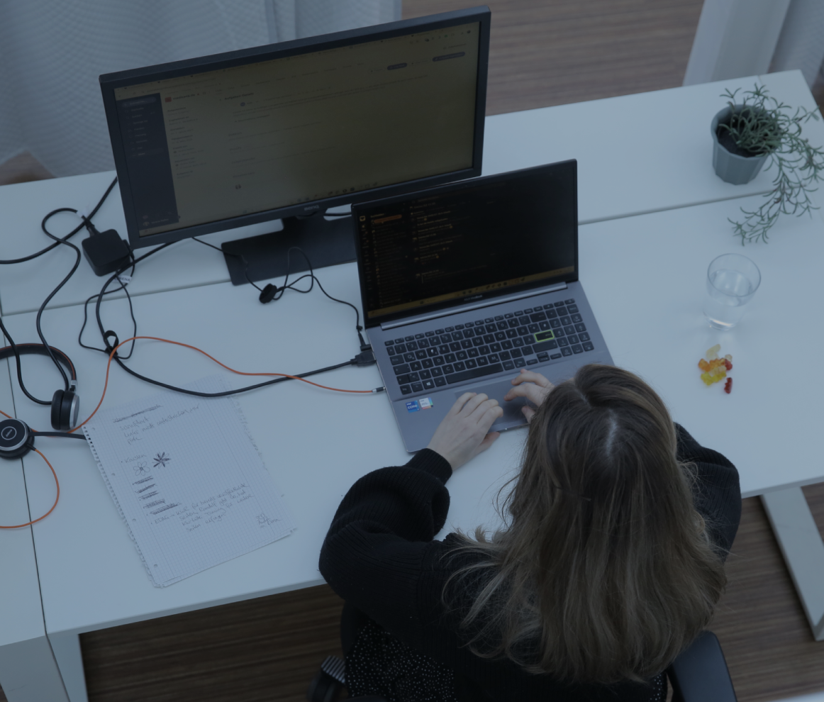 Person tippt an einem Laptop am Schreibtisch im Büro, aufgenommen aus der Vogelperspektive, mit externem Monitor und kleiner Pflanze