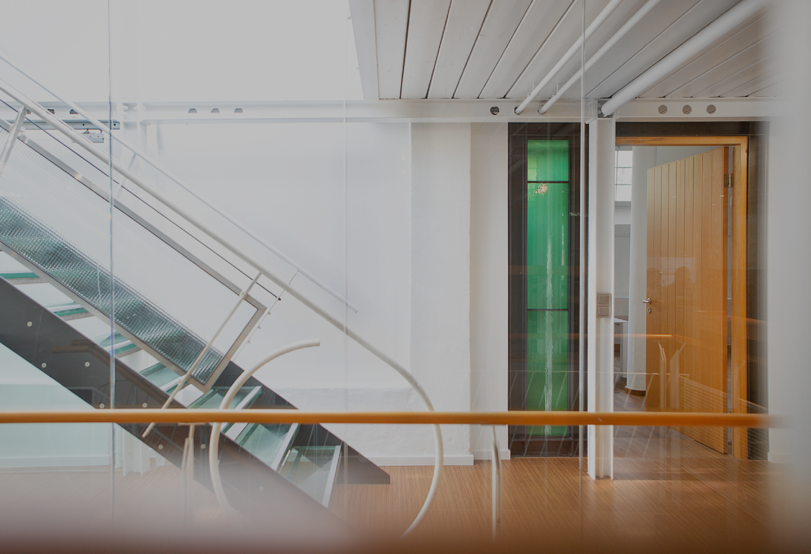 Heller Büroflur mit moderner Glas- und Metalltreppe, weißen Wänden und Holzboden; rechts ein offener Raum mit Holztür und grünem Glaselement, klare, minimalistische Architektur