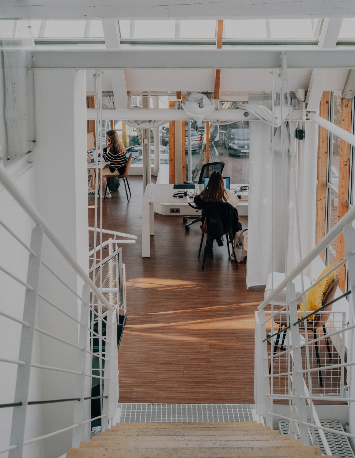 Blick von einer Treppe in ein helles, modernes Büro mit Holzfußboden, Glasfronten und zwei Personen, die an Schreibtischen arbeiten.