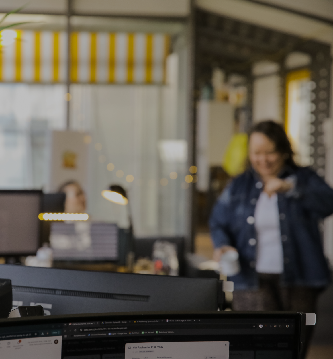 Unscharfe Büroszene mit Monitor im Vordergrund und einer Person, die im Hintergrund mit einer Tasse durch das Büro geht