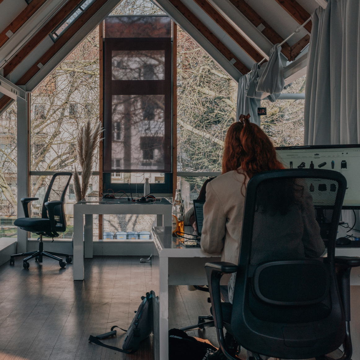 Zwei Personen arbeiten an separaten Schreibtischen in einem hellen Büro mit großen Fenstern, sichtbaren Holzbalken und Blick ins Grüne