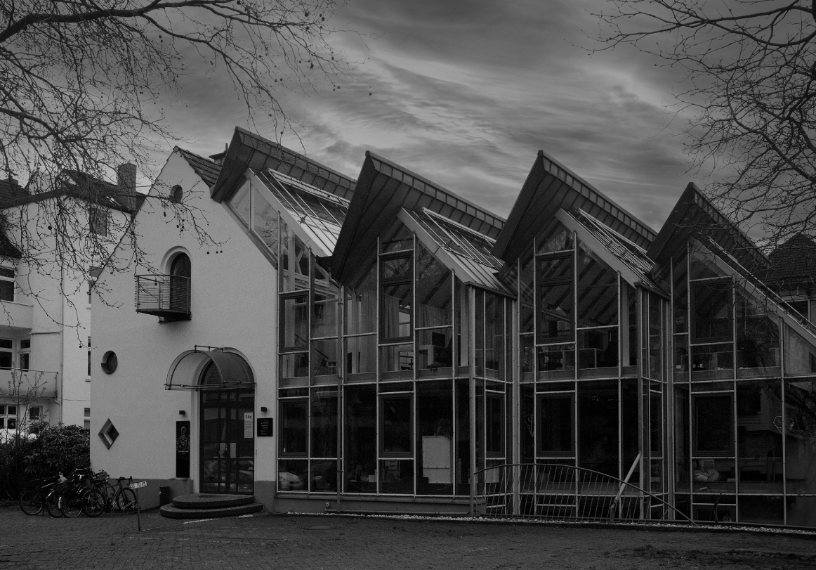 Schwarzweißaufnahme eines modernen Bürogebäudes mit großer Glasfassade und markanten, schräg auskragenden Dächern; davor Bäume ohne Blätter, ein gepflasterter Vorplatz