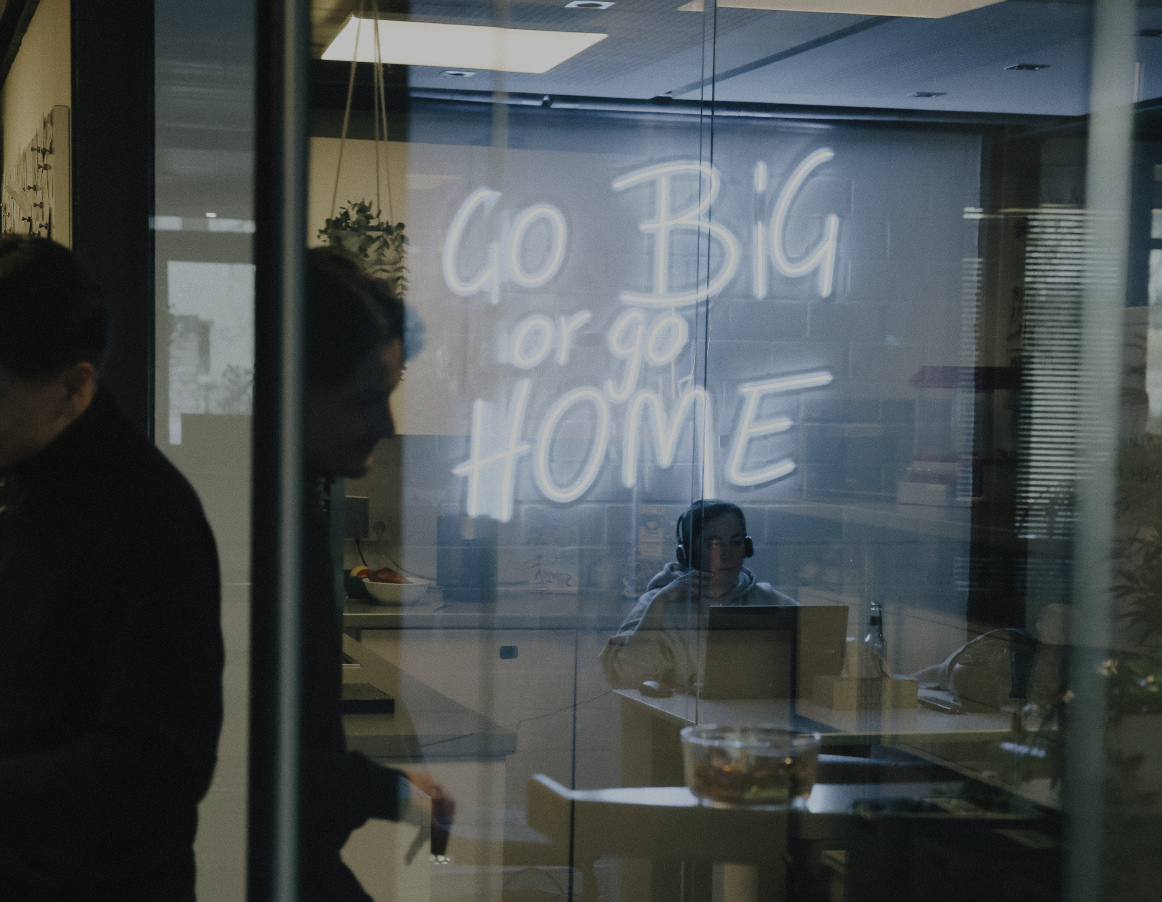 Blick durch eine Glaswand in einen Besprechungsraum mit leuchtender Neon-Schrift „Go Big or Go Home“ und arbeitenden Personen