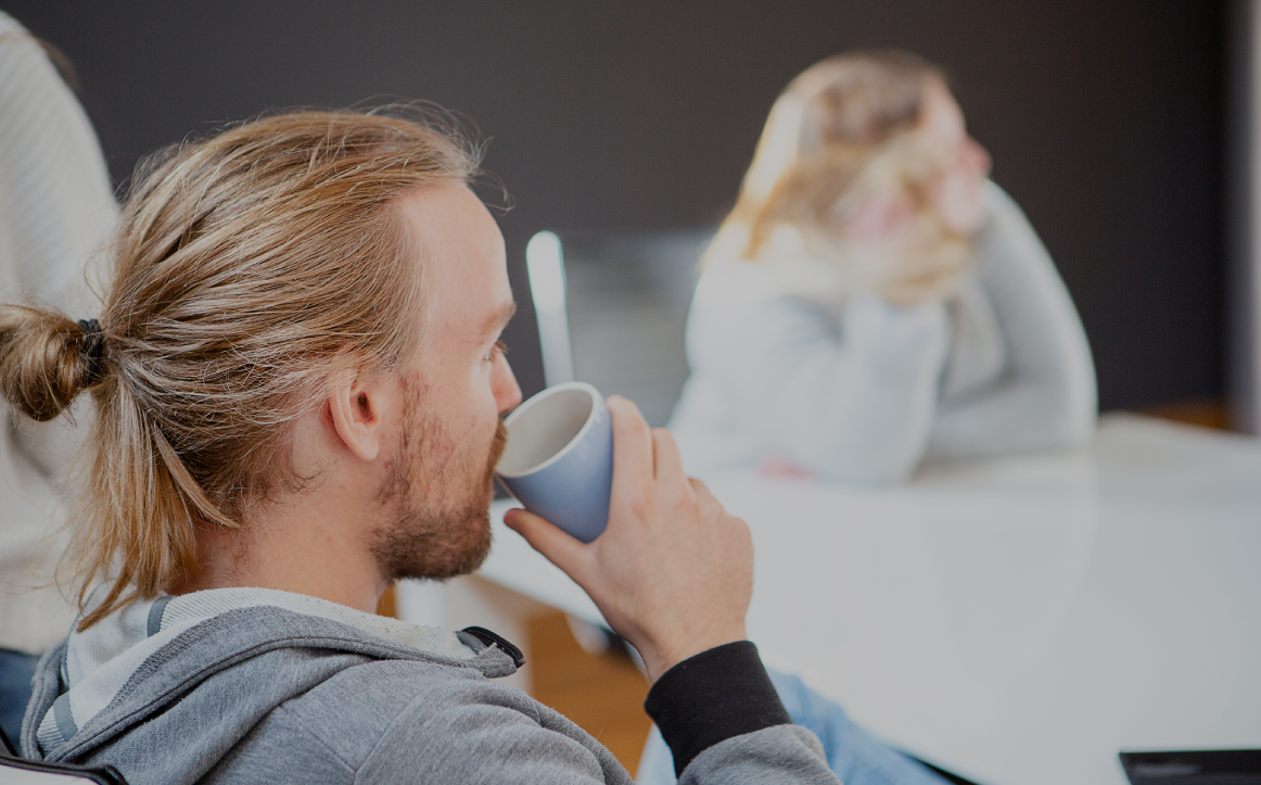 Person mit zusammengebundenem Haar trinkt aus einer Tasse in einem Besprechungsraum; im unscharfen Hintergrund sitzt eine weitere Person am Tisch, helle, entspannte Büroszene