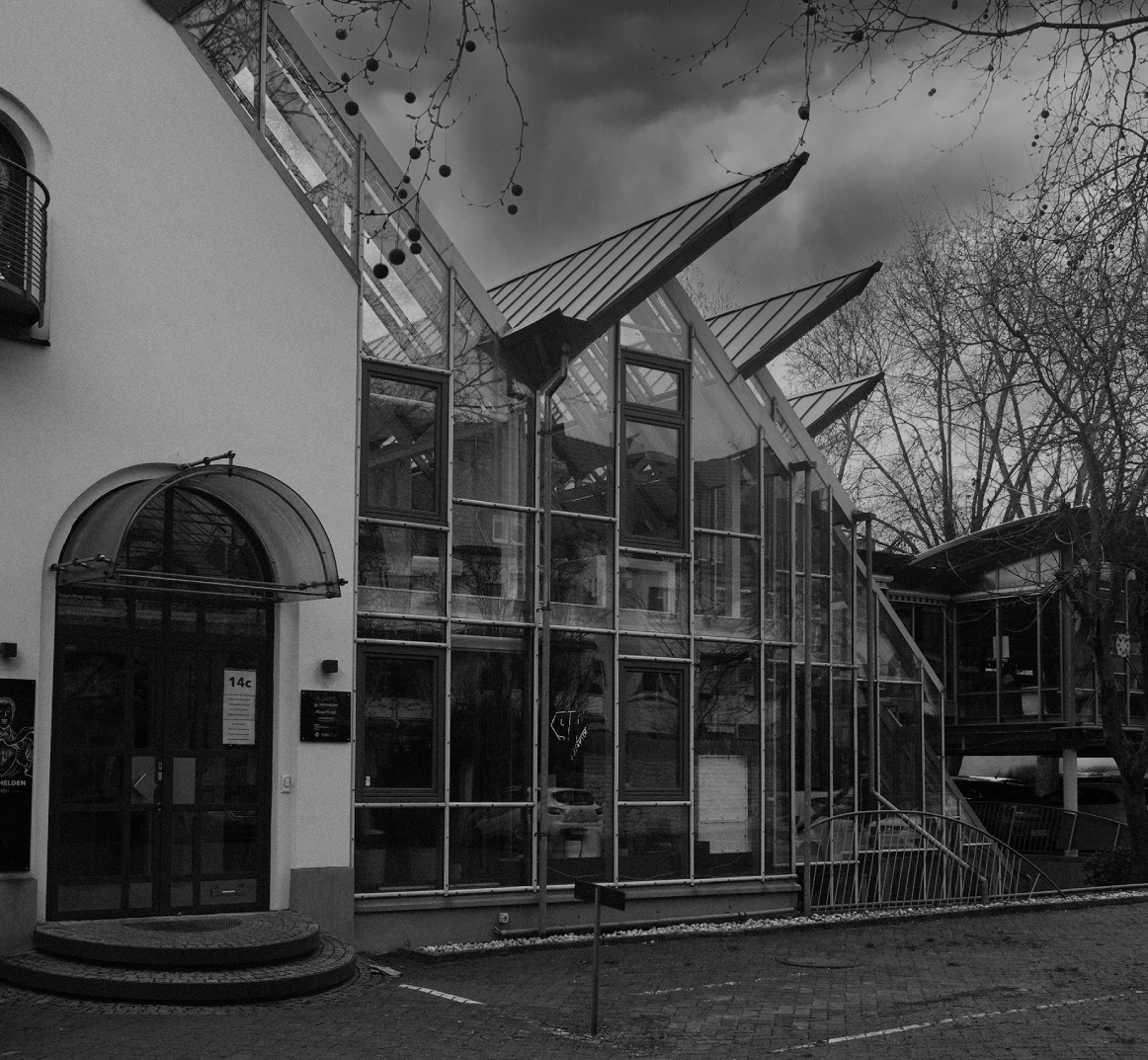 Schwarzweißaufnahme eines modernen Bürogebäudes mit großer Glasfassade und markanten, schräg auskragenden Dächern; davor Bäume ohne Blätter, ein gepflasterter Vorplatz und parkende Autos