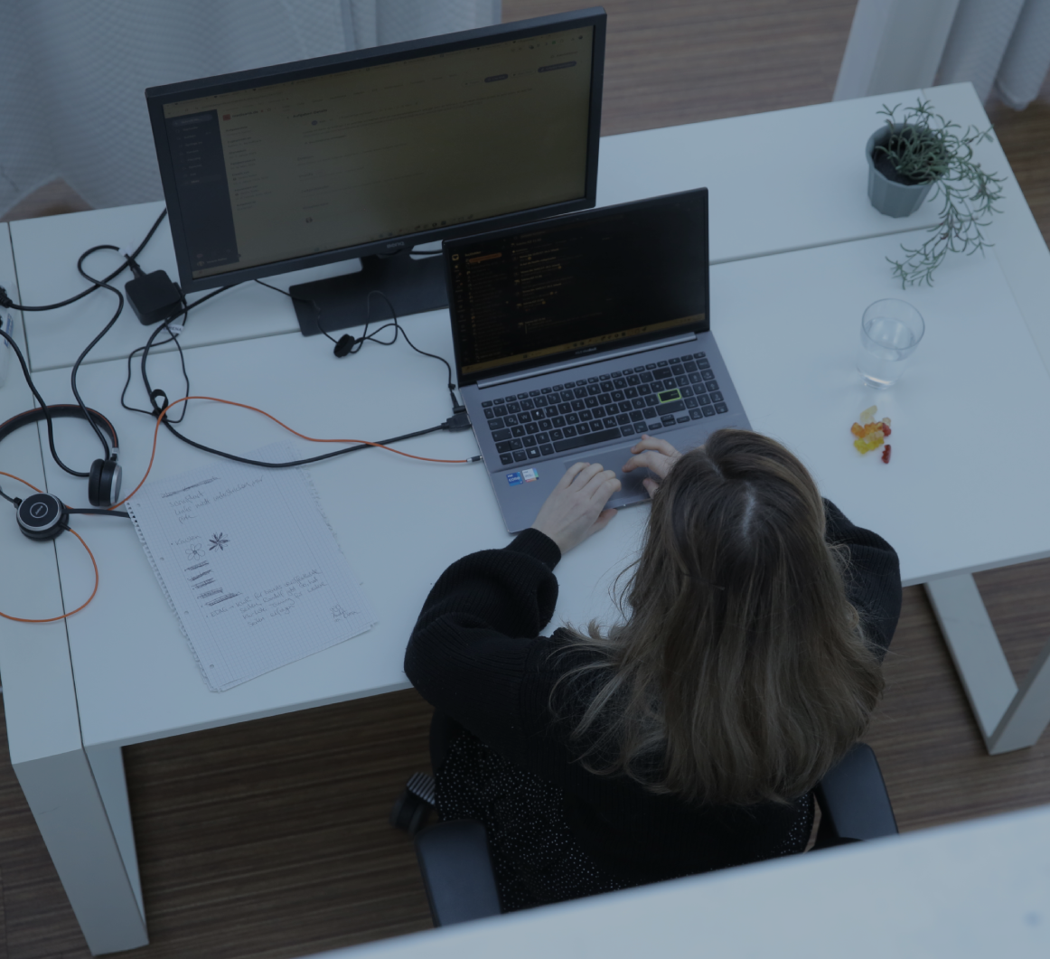 Person tippt an einem Laptop am Schreibtisch im Büro, aufgenommen aus der Vogelperspektive, mit externem Monitor und kleiner Pflanze