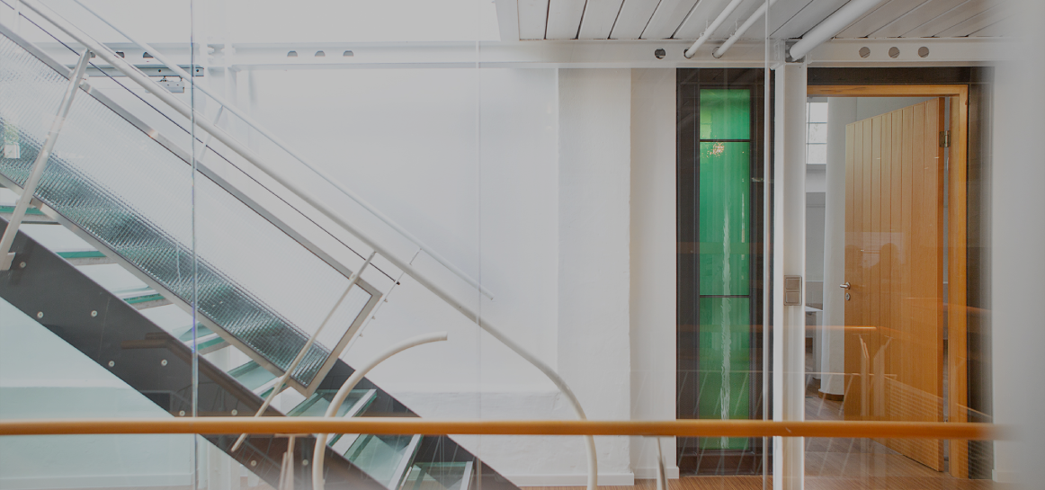 Heller Büroflur mit moderner Glas- und Metalltreppe, weißen Wänden und Holzboden; rechts ein offener Raum mit Holztür und grünem Glaselement, klare, minimalistische Architektur