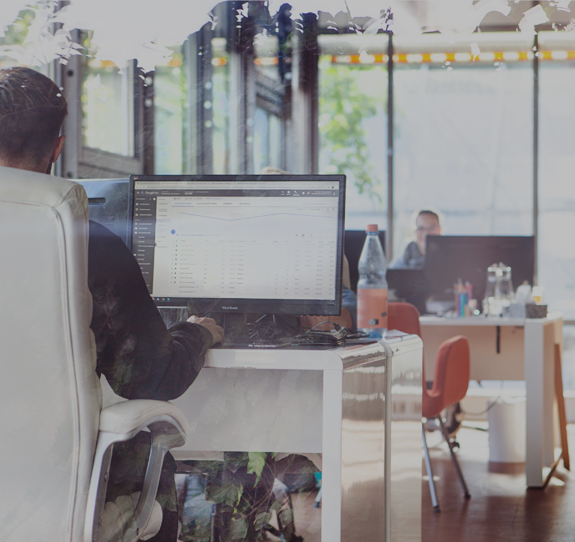 Heller Büroraum mit mehreren Arbeitsplätzen_ Im Vordergrund sitzt eine Person am Schreibtisch vor einem großen Monitor mit Tabellenansicht, im Hintergrund arbeiten weitere Personen an Laptops