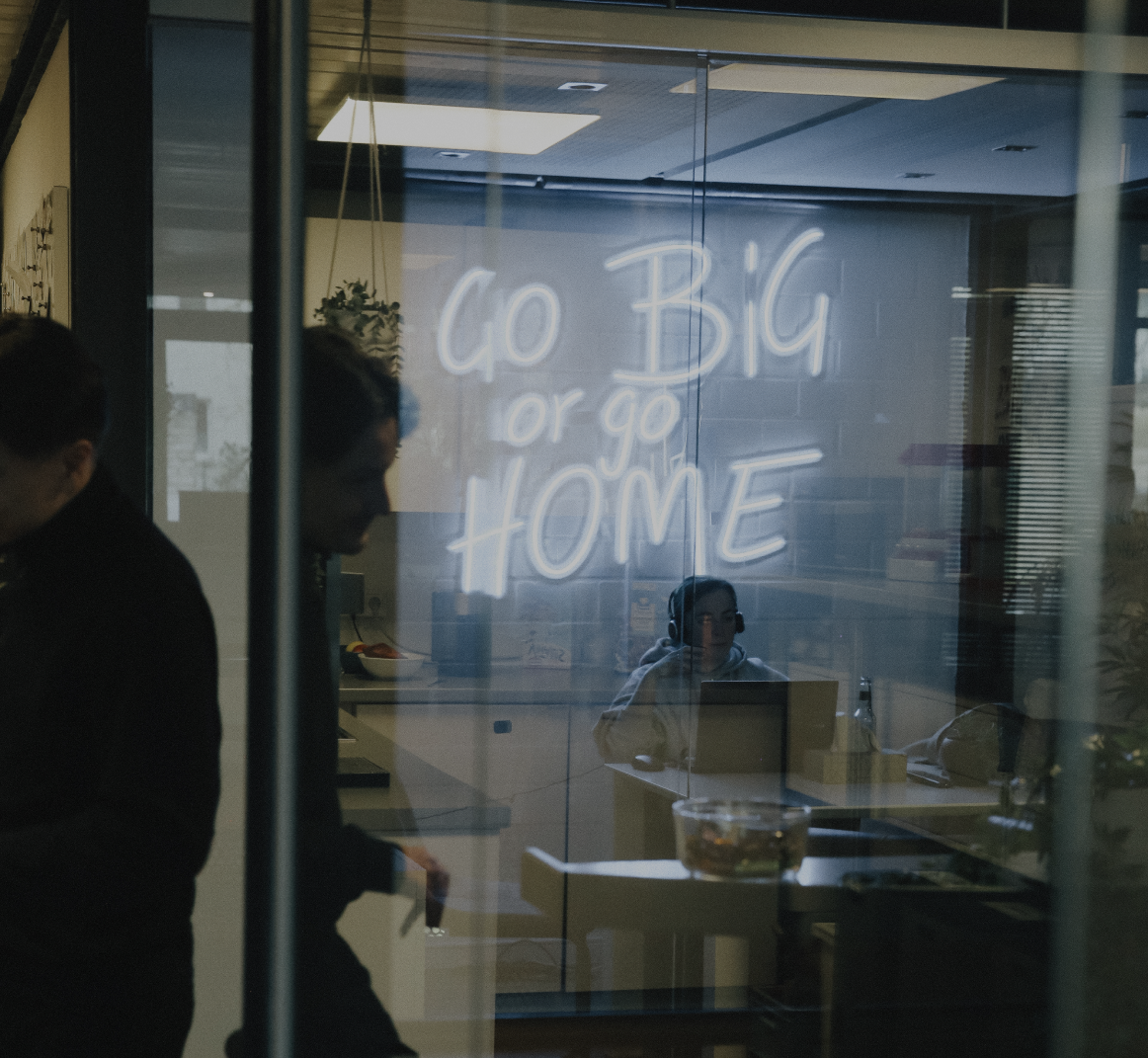 Blick durch eine Glaswand in einen Besprechungsraum mit leuchtender Neon-Schrift „Go Big or Go Home“ und arbeitenden Personen