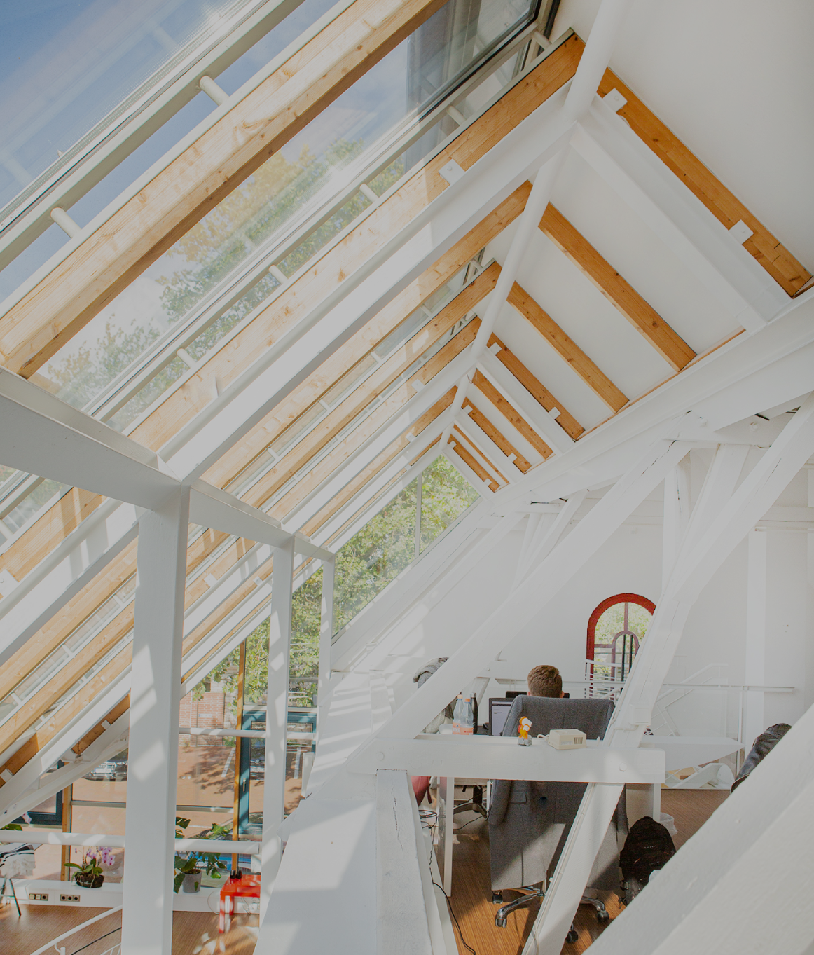 Helles Dachgeschossbüro mit großen Glasdachfenstern und sichtbaren Holz- und Stahlträgern; eine Person sitzt an einem Schreibtisch, viel Tageslicht fällt in den offenen, modernen Arbeitsbereich