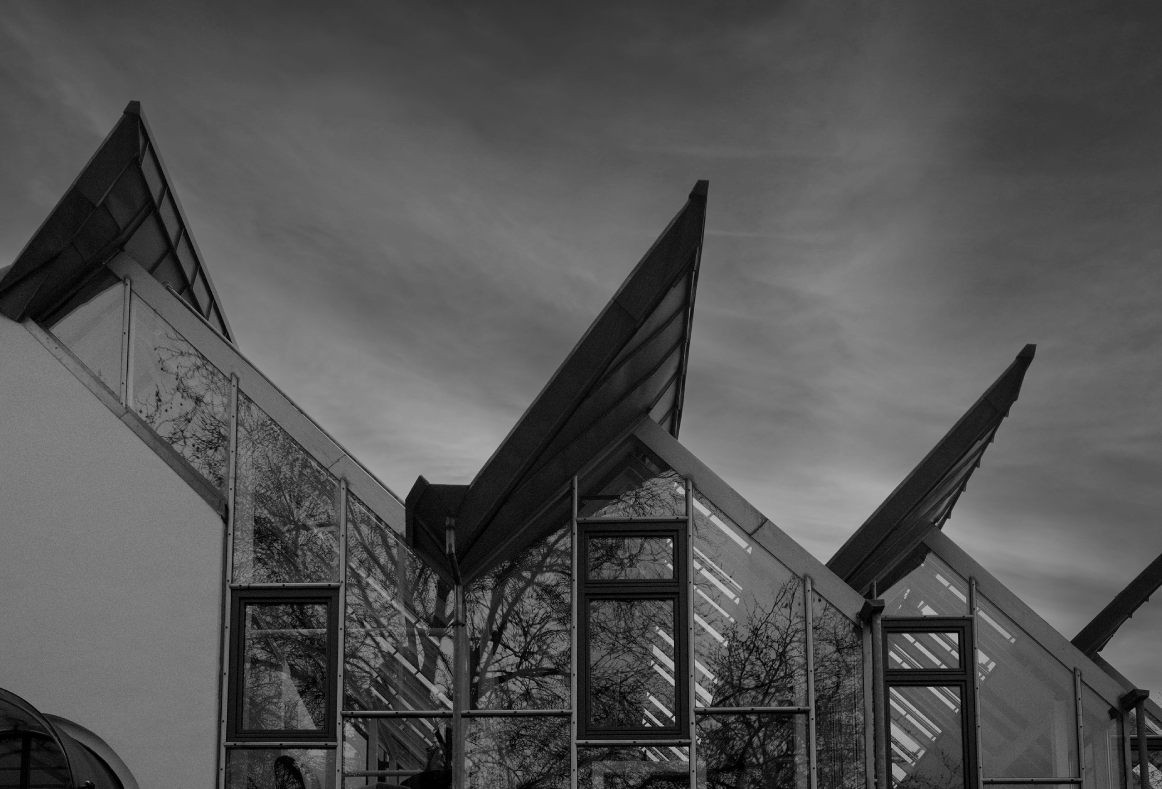 Schwarzweißaufnahme der Glasfassade eines modernen Bürogebäudes mit mehreren spitz zulaufenden, schräg auskragenden Dachkonstruktionen; Spiegelungen von kahlen Bäumen in den Fenstern und ein bewölkter