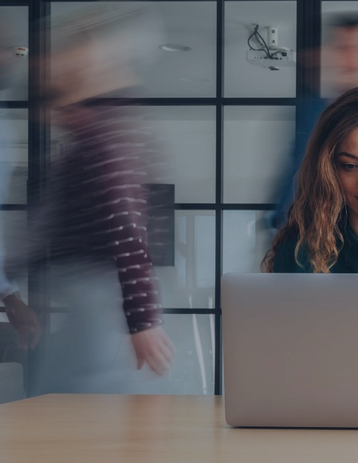 Frau arbeitet am Laptop in einem Büro, im Hintergrund gehen unscharf Personen vorbei.