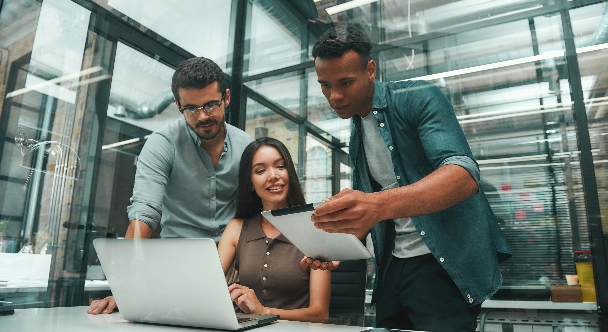 Eine Gruppe von drei jungen und positiven Mitarbeitern nutzt moderne Technologien und unterhält sich während der Arbeit in einem modernen Büro.