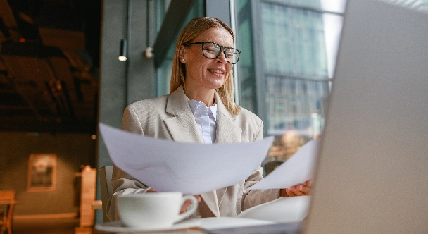 Eine smarte Geschäftsfrau mit Brille arbeitet in einem Café an Dokumenten und ihrem Laptop.