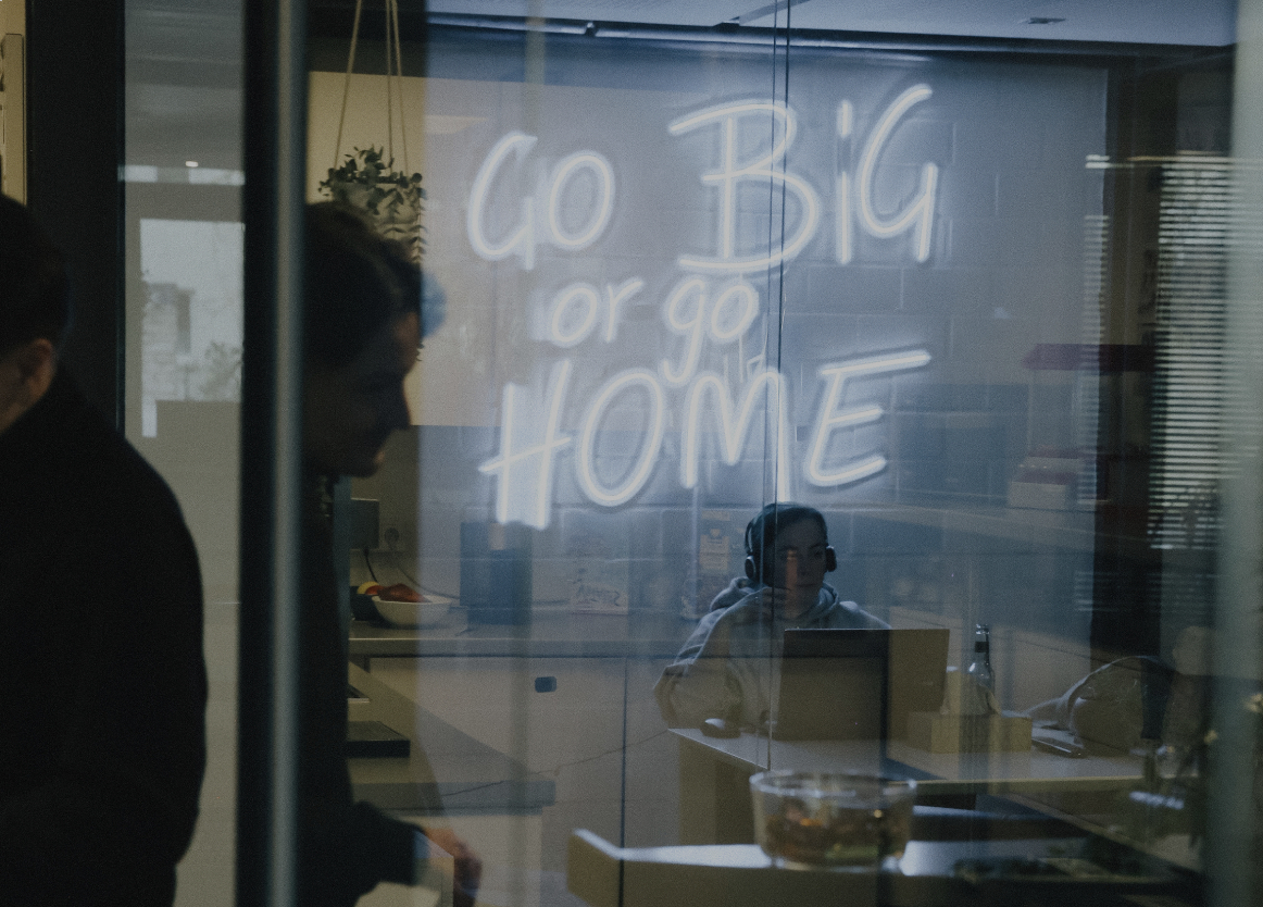 Blick durch eine Glaswand in einen Besprechungsraum mit leuchtender Neon-Schrift „Go Big or Go Home“ und arbeitenden Personen