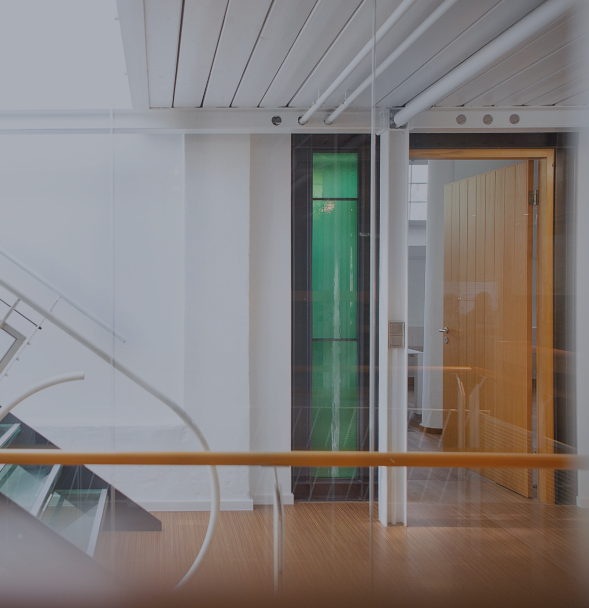 Heller Büroflur mit moderner Glas- und Metalltreppe, weißen Wänden und Holzboden; rechts ein offener Raum mit Holztür und grünem Glaselement, klare, minimalistische Architektur
