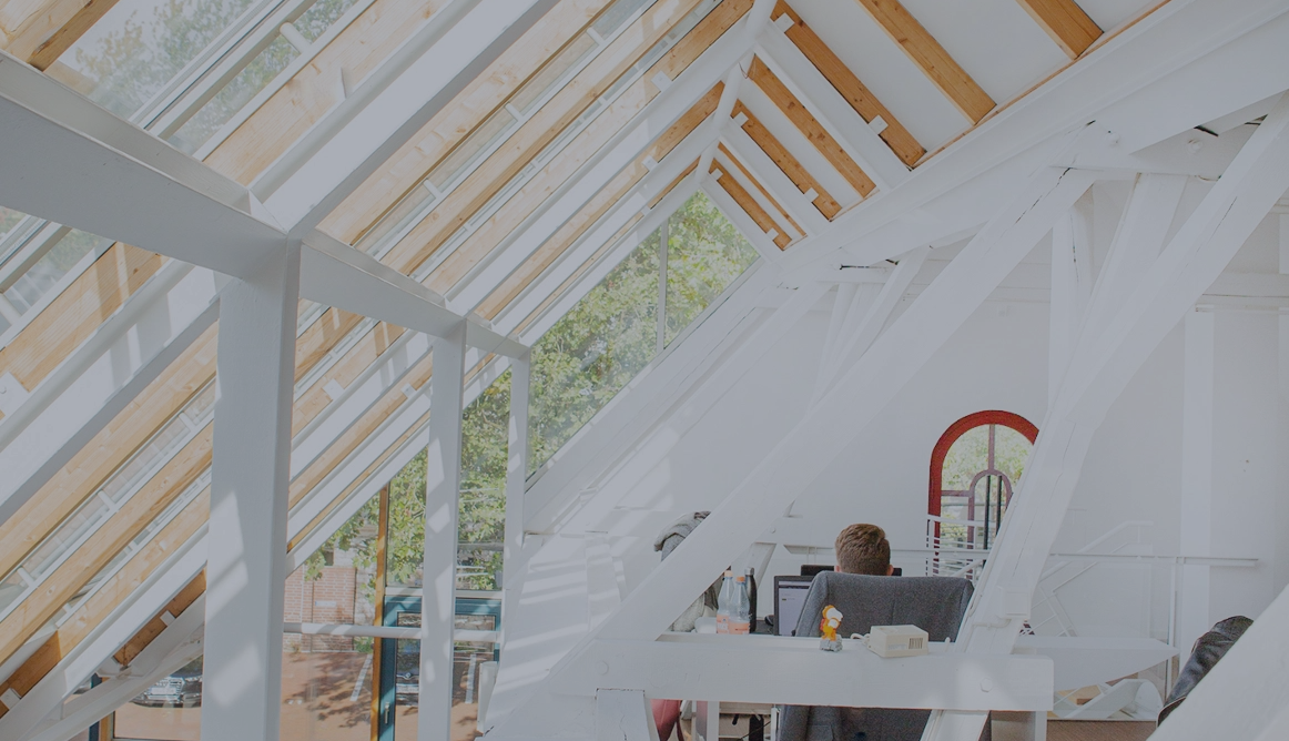 Helles Dachgeschossbüro mit großen Glasdachfenstern und sichtbaren Holz- und Stahlträgern; eine Person sitzt an einem Schreibtisch, viel Tageslicht fällt in den offenen, modernen Arbeitsbereich