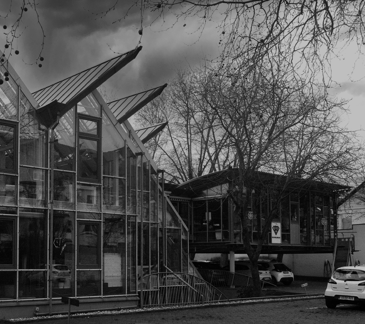 Schwarzweißaufnahme eines modernen Bürogebäudes mit großer Glasfassade und markanten, schräg auskragenden Dächern; davor Bäume ohne Blätter, ein gepflasterter Vorplatz und parkende Autos