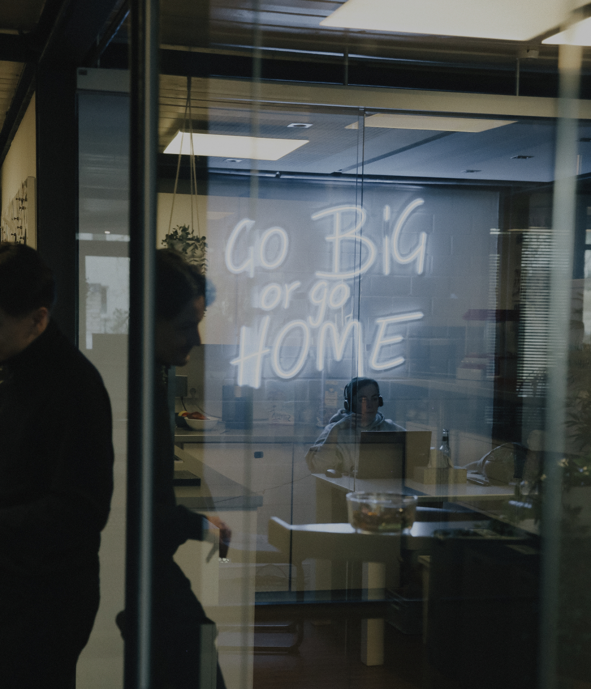 Blick durch eine Glaswand in einen Besprechungsraum mit leuchtender Neon-Schrift „Go Big or Go Home“ und arbeitenden Personen