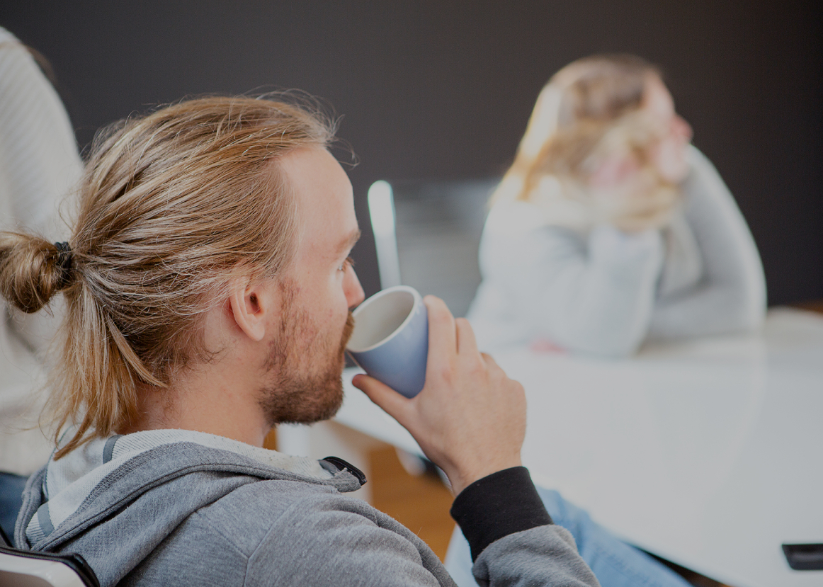Person mit zusammengebundenem Haar trinkt aus einer Tasse in einem Besprechungsraum; im unscharfen Hintergrund sitzt eine weitere Person am Tisch, helle, entspannte Büroszene