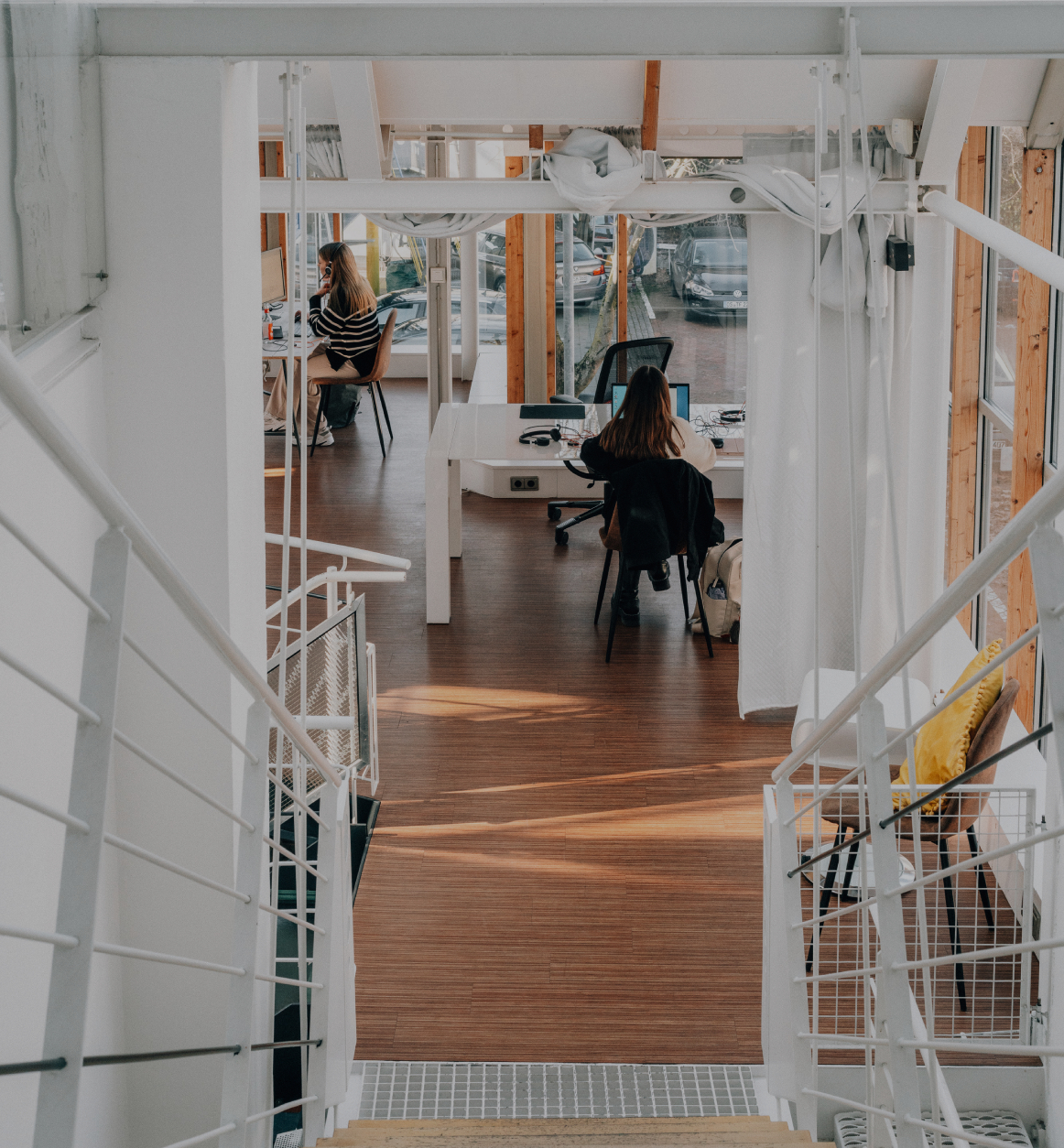 Blick von einer Treppe in ein helles, modernes Büro mit Holzfußboden, Glasfronten und zwei Personen, die an Schreibtischen arbeiten.