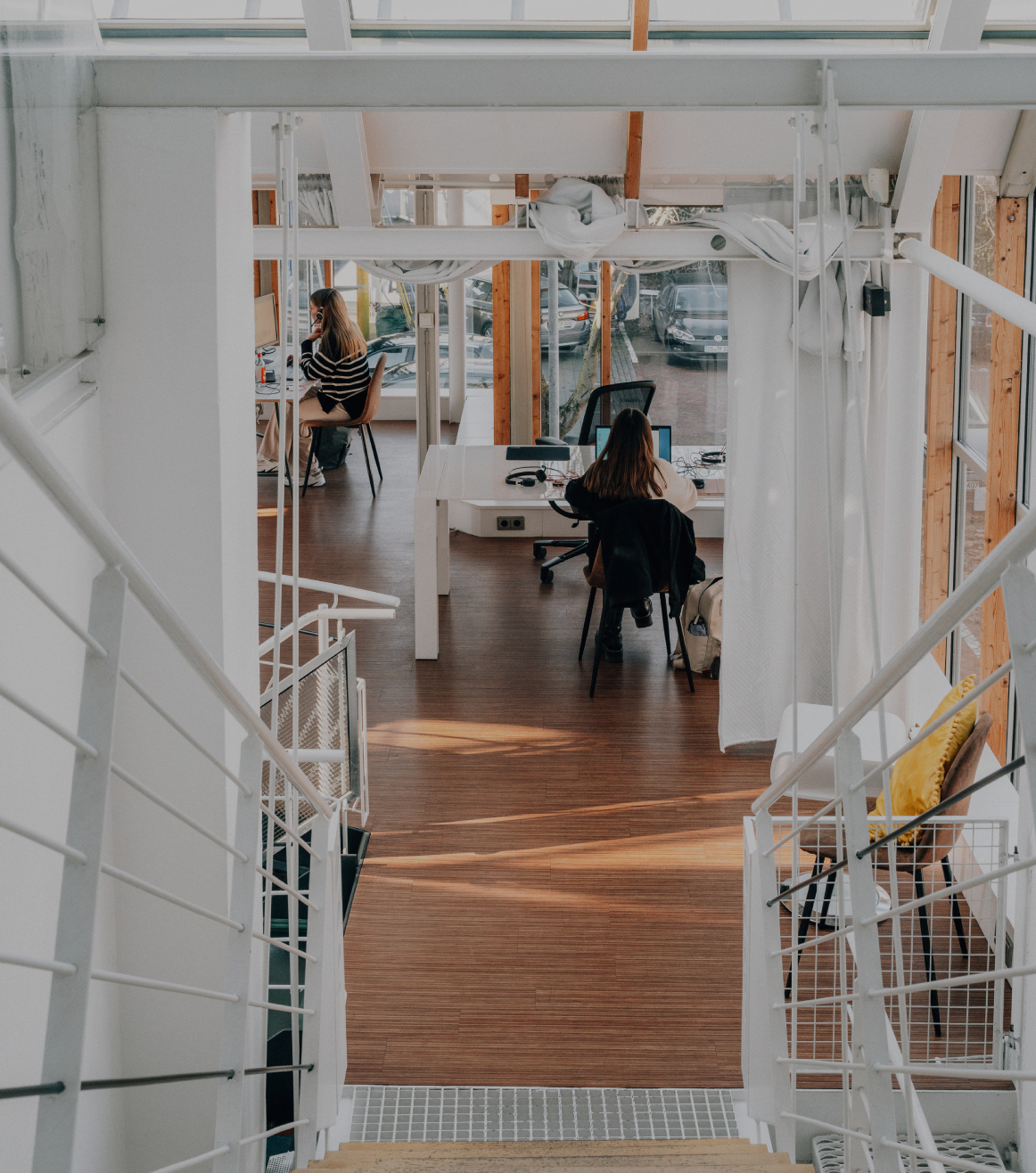 Blick von einer Treppe in ein helles, modernes Büro mit Holzfußboden, Glasfronten und zwei Personen, die an Schreibtischen arbeiten.