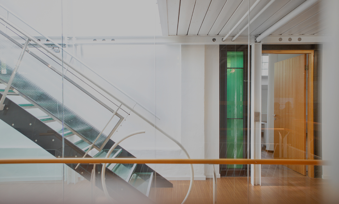 Heller Büroflur mit moderner Glas- und Metalltreppe, weißen Wänden und Holzboden; rechts ein offener Raum mit Holztür und grünem Glaselement, klare, minimalistische Architektur