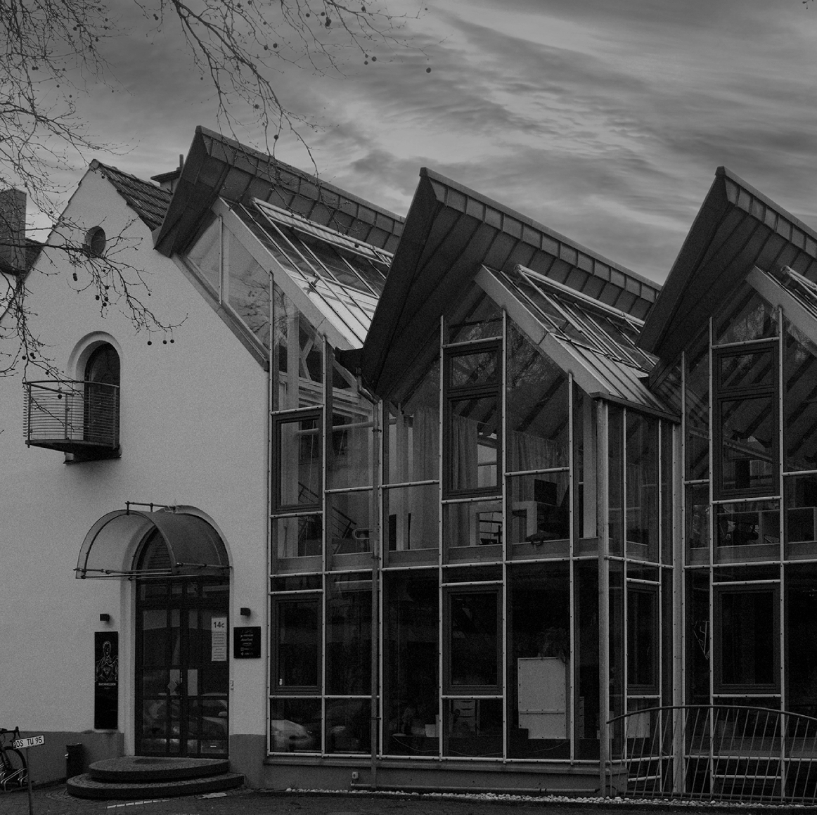 Schwarzweißaufnahme eines modernen Bürogebäudes mit großer Glasfassade und markanten, schräg auskragenden Dächern; davor Bäume ohne Blätter, ein gepflasterter Vorplatz