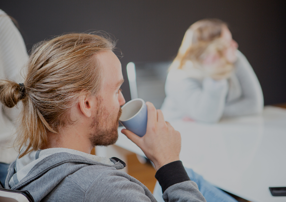 Person mit zusammengebundenem Haar trinkt aus einer Tasse in einem Besprechungsraum; im unscharfen Hintergrund sitzt eine weitere Person am Tisch, helle, entspannte Büroszene