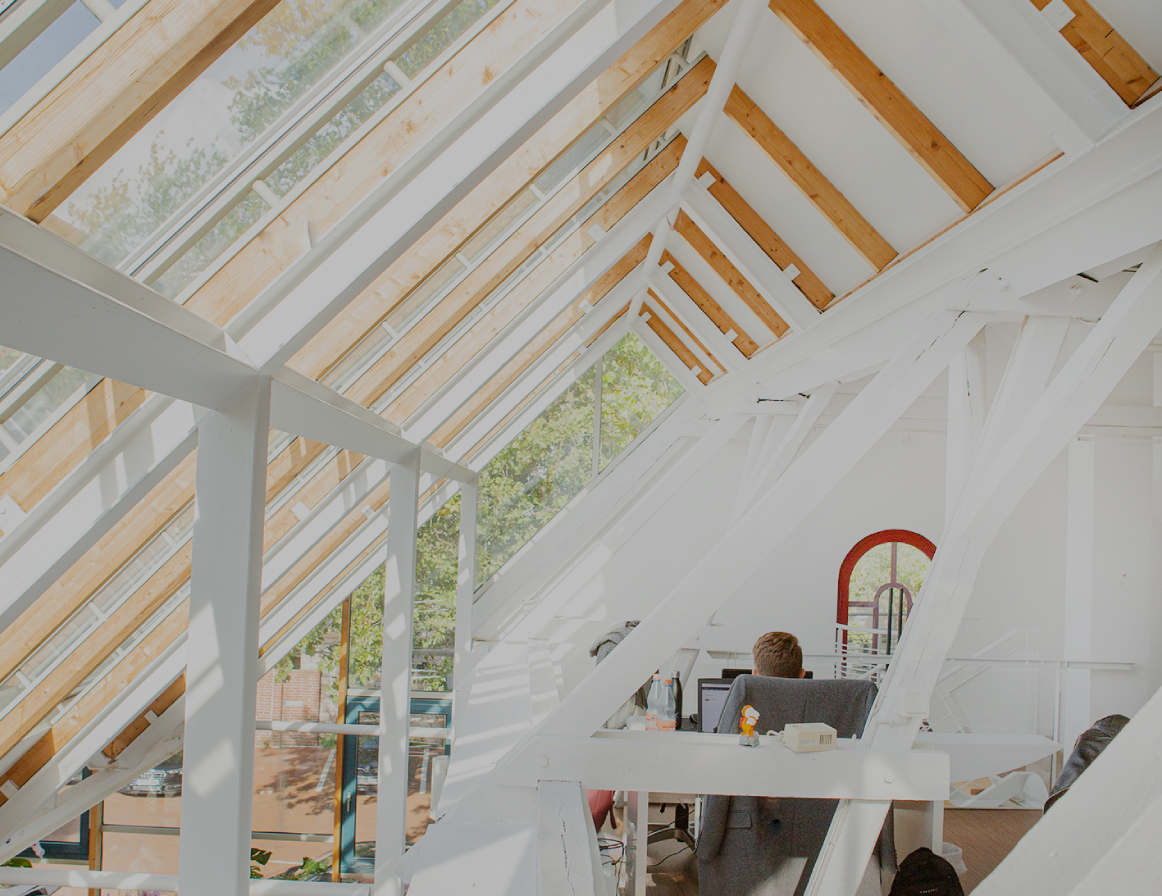 Helles Dachgeschossbüro mit großen Glasdachfenstern und sichtbaren Holz- und Stahlträgern; eine Person sitzt an einem Schreibtisch, viel Tageslicht fällt in den offenen, modernen Arbeitsbereich