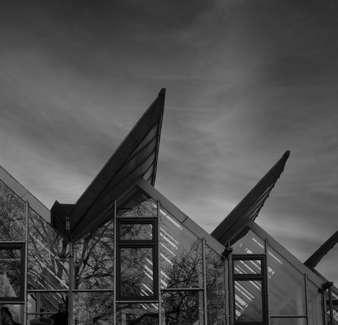 Schwarzweißaufnahme der Glasfassade eines modernen Bürogebäudes mit mehreren spitz zulaufenden, schräg auskragenden Dachkonstruktionen; Spiegelungen von kahlen Bäumen in den Fenstern und ein bewölkter Himmel im Hinterg