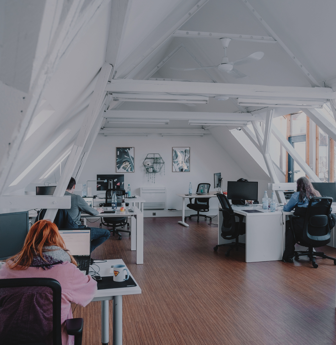 Großzügiges, helles Büro mit schrägen Dachbalken, mehreren Arbeitsplätzen und Personen, die an Schreibtischen arbeiten