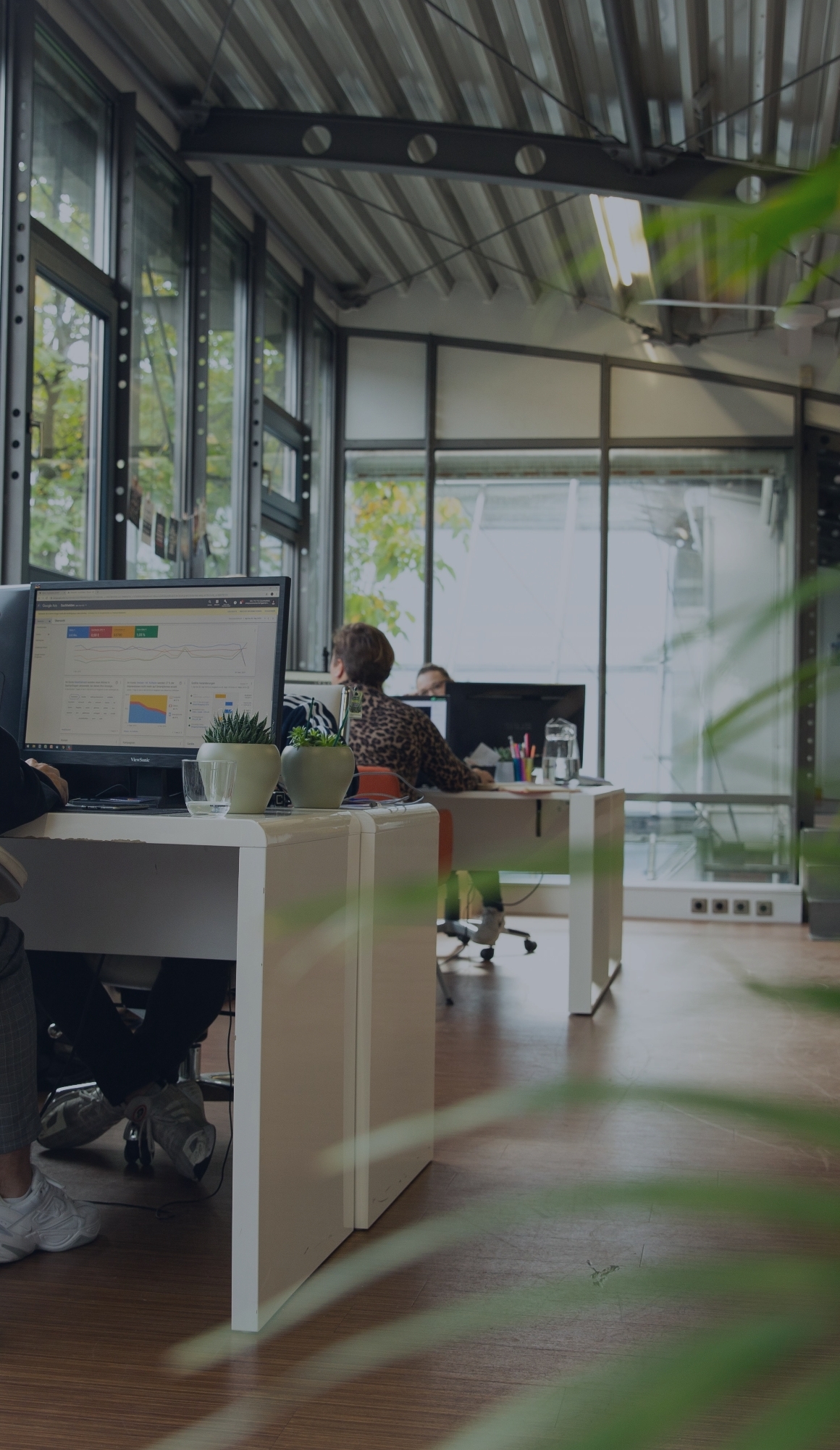 Personen arbeiten an Schreibtischen in einem hellen Büro, im Vordergrund ein Monitor mit Diagrammen und kleine Pflanzen.