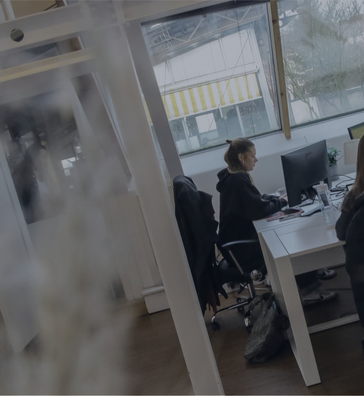 Person arbeitet an einem Schreibtisch im Büro, seitlich durch eine Glaswand fotografiert, mit Fensterfront im Hintergrund.