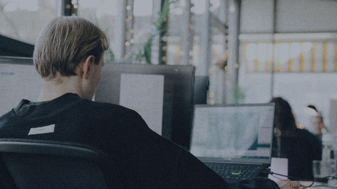 Person sitzt an einem Schreibtisch im Büro und arbeitet an einem Computer mit mehreren Bildschirmen. 