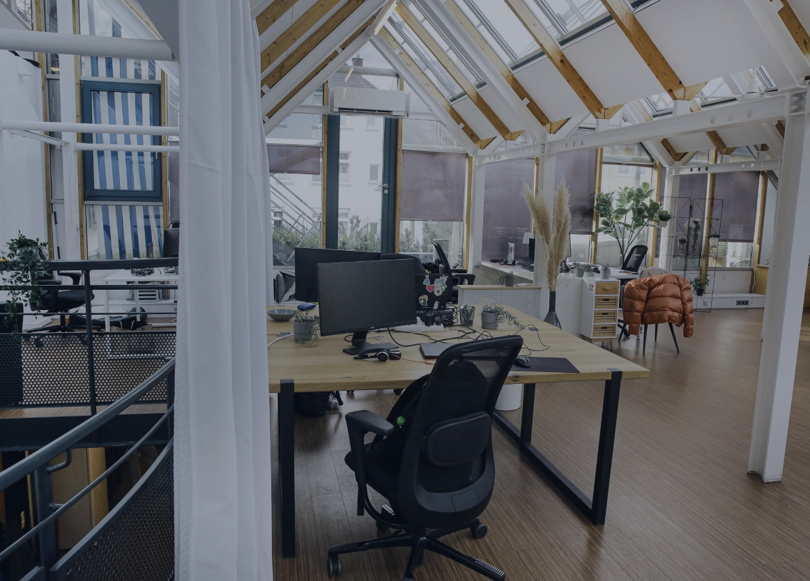 Heller, moderner Büroraum mit Schreibtischen, Monitoren und viel Tageslicht unter einem Glasdach.