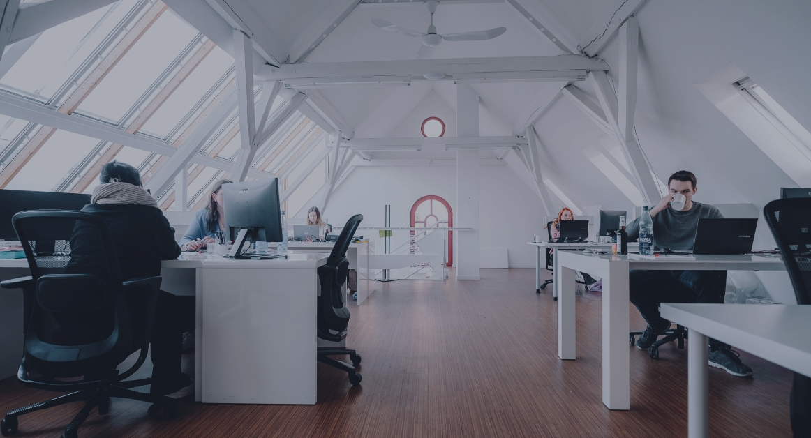 Helles Großraumbüro mit mehreren Personen an Schreibtischen unter einem Dach mit großen Fenstern.