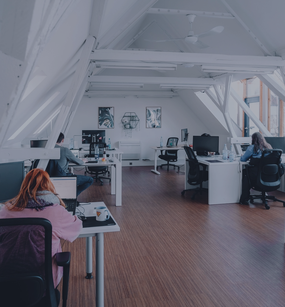 Großzügiges, helles Büro mit schrägen Dachbalken, mehreren Arbeitsplätzen und Personen, die an Schreibtischen arbeiten