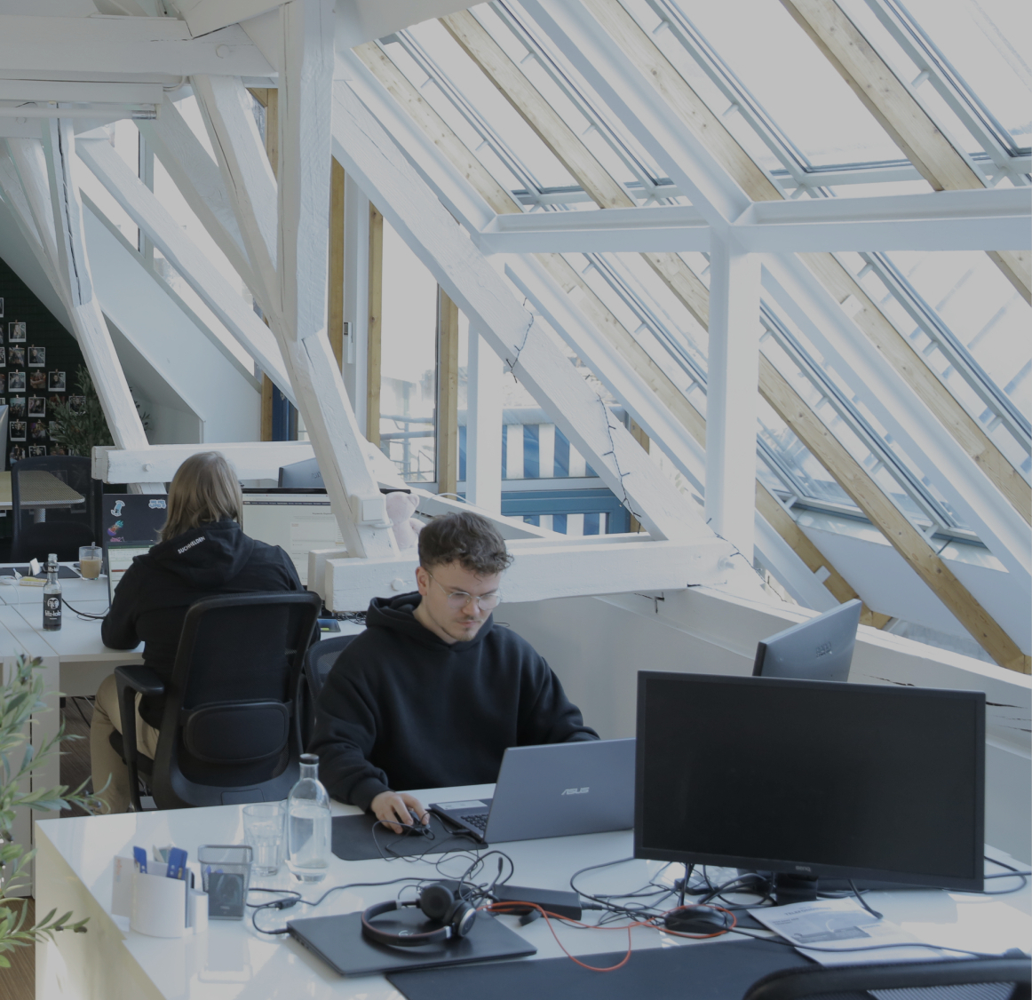 Mehrere Personen arbeiten an Schreibtischen in einem offenen Büro mit schrägen Dachfenstern, Pflanzen und einer beleuchteten „#Suchhelden“-Wand im Hintergrund