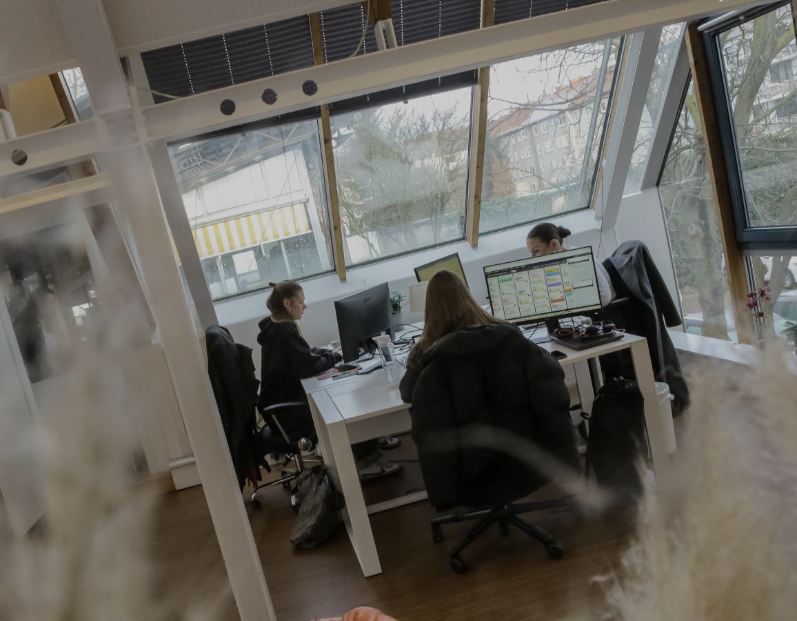 Person arbeitet an einem Schreibtisch im Büro, seitlich durch eine Glaswand fotografiert, mit Fensterfront im Hintergrund.