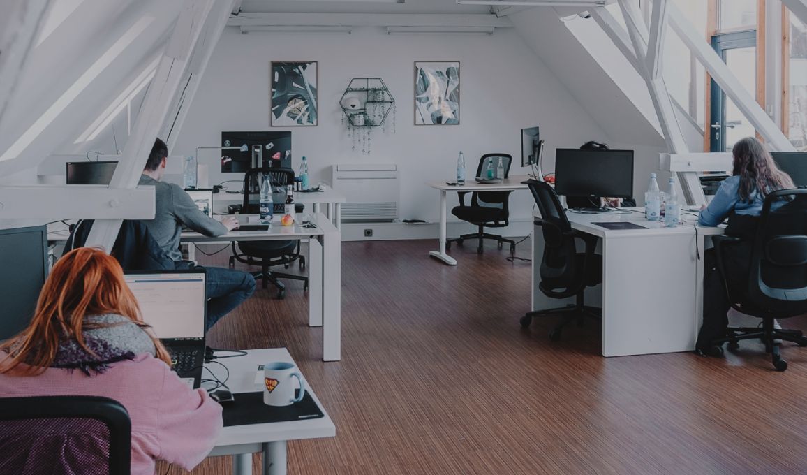 Großzügiges, helles Büro mit schrägen Dachbalken, mehreren Arbeitsplätzen und Personen, die an Schreibtischen arbeiten