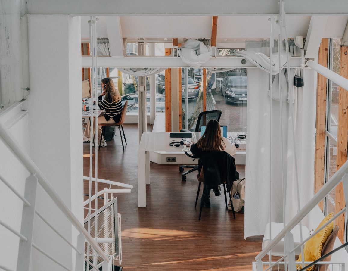 Blick von einer Treppe in ein helles, modernes Büro mit Holzfußboden, Glasfronten und zwei Personen, die an Schreibtischen arbeiten.